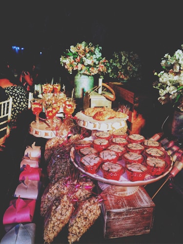 A beautifully decorated dessert table featuring a variety of treats, including cupcakes with cream tops placed on a stand, wrapped bags of popcorn, and multiple decorative gift boxes. Several glasses of a vibrant pink beverage are also visible, along with lush floral arrangements in vases enhancing the rich, celebratory atmosphere.
