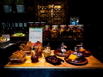 A table with a variety of foods including cheeses under glass cloches, jars of pickled vegetables, a plate of crispbread, and small dishes of nuts and fruits. In the background, jars of pickles and a decorative black panel are visible. The lighting creates a warm and inviting atmosphere.