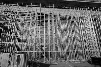 A dense grid of scaffolding frames the entire view, creating a complex, geometric pattern. The scene appears to be part of a large construction or renovation project. The scaffolding stretches vertically and horizontally, forming a tunnel-like structure. There is minimal lighting, casting shadows and highlights on the metallic surfaces.