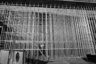 A dense grid of scaffolding frames the entire view, creating a complex, geometric pattern. The scene appears to be part of a large construction or renovation project. The scaffolding stretches vertically and horizontally, forming a tunnel-like structure. There is minimal lighting, casting shadows and highlights on the metallic surfaces.