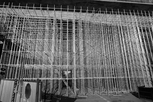 A dense grid of scaffolding frames the entire view, creating a complex, geometric pattern. The scene appears to be part of a large construction or renovation project. The scaffolding stretches vertically and horizontally, forming a tunnel-like structure. There is minimal lighting, casting shadows and highlights on the metallic surfaces.