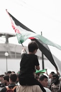 A group of people gathered with one individual sitting on another's shoulders, holding a large flag with black, white, green, and red sections. The atmosphere appears focused and determined, with multiple people looking up towards the flag.