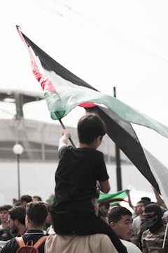 A group of people gathered with one individual sitting on another's shoulders, holding a large flag with black, white, green, and red sections. The atmosphere appears focused and determined, with multiple people looking up towards the flag.