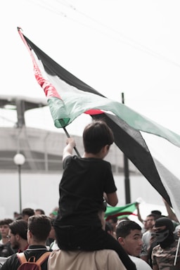 A group of people gathered with one individual sitting on another's shoulders, holding a large flag with black, white, green, and red sections. The atmosphere appears focused and determined, with multiple people looking up towards the flag.