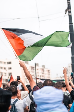 A crowd of people is gathered, many holding up mobile phones. A prominent flag is being waved above the group, with colors red, white, black, and green. The background shows a few buildings.