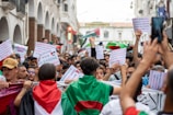 A large group of people gathered in an urban setting, holding various signs and banners related to a political demonstration. Many participants wear flags over their shoulders, and signs contain messages of solidarity and protest. The scene is lively with a diverse crowd, set against a backdrop of arched white buildings.