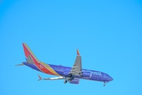 A commercial airplane is in flight against a clear blue sky, with visible landing gear and a tail featuring vibrant colors and branding.