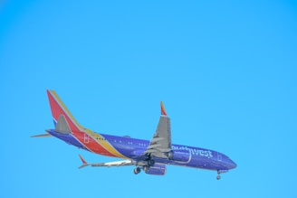 A commercial airplane is in flight against a clear blue sky, with visible landing gear and a tail featuring vibrant colors and branding.