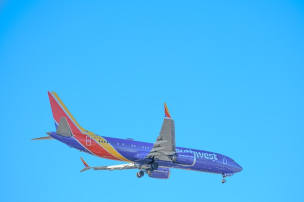 A commercial airplane is in flight against a clear blue sky, with visible landing gear and a tail featuring vibrant colors and branding.