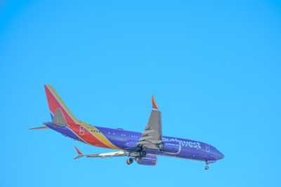 A commercial airplane is in flight against a clear blue sky, with visible landing gear and a tail featuring vibrant colors and branding.