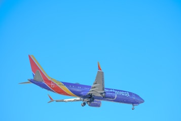 A commercial airplane is in flight against a clear blue sky, with visible landing gear and a tail featuring vibrant colors and branding.