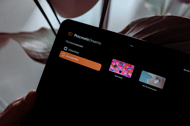 A digital tablet screen displays the Procreate Dreams application. The interface shows options like iCloud Drive and a section 'на моем iPad'. Two thumbnail images are visible, each representing a different artistic project: 'Spark dream' with colorful abstract art, and 'Into The Future dream' featuring an illustration of a character's profile. The background features leaves, suggesting a natural or indoor plant setting.