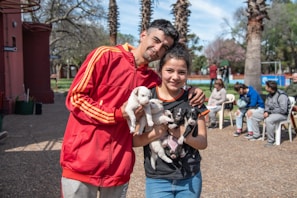 Two people are happily holding four puppies in an outdoor park setting. They are standing close together, with one person wearing a red jacket and the other in a black shirt. In the background, several people are sitting on chairs surrounded by trees and some buildings, creating a relaxed, communal atmosphere.