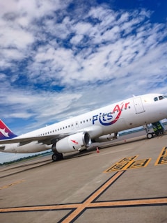 A commercial airplane is on the tarmac under a partly cloudy sky. The aircraft bears the logo 'Fits Air' on its fuselage, and there are a few orange traffic cones placed nearby. The runway has visible markings, and there are patches of greenery seen in the background.
