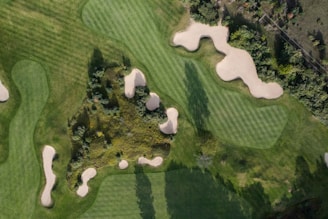 An aerial view of a golf course featuring lush green fairways with neatly cut grass patterns and several sand bunkers scattered throughout. Dense patches of vegetation and trees surround the golfing area, adding a natural backdrop to the setting.