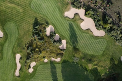 An aerial view of a golf course featuring lush green fairways with neatly cut grass patterns and several sand bunkers scattered throughout. Dense patches of vegetation and trees surround the golfing area, adding a natural backdrop to the setting.