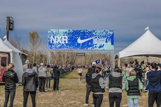 A cross-country race event with spectators lined along the sides of a dirt path under a blue and white banner with the text 'NXR' and 'Nike Cross Regionals'. The scene is surrounded by tents and spectators wearing winter clothing, indicating a chilly outdoor setting. One runner is visible approaching the finish line.