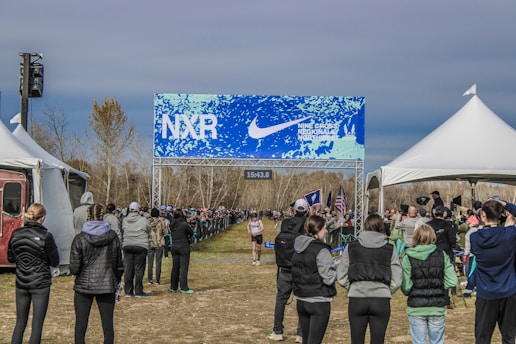 A cross-country race event with spectators lined along the sides of a dirt path under a blue and white banner with the text 'NXR' and 'Nike Cross Regionals'. The scene is surrounded by tents and spectators wearing winter clothing, indicating a chilly outdoor setting. One runner is visible approaching the finish line.