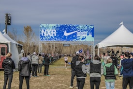 A cross-country race event with spectators lined along the sides of a dirt path under a blue and white banner with the text 'NXR' and 'Nike Cross Regionals'. The scene is surrounded by tents and spectators wearing winter clothing, indicating a chilly outdoor setting. One runner is visible approaching the finish line.