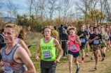 A group of young athletes is actively participating in a cross-country race on a natural, grassy course surrounded by trees. The runners are wearing numbered bibs for identification and are dressed in various athletic gear appropriate for the weather. The background features bare trees and sparse foliage, indicating a likely autumn setting.