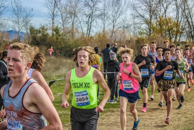 A group of young athletes is actively participating in a cross-country race on a natural, grassy course surrounded by trees. The runners are wearing numbered bibs for identification and are dressed in various athletic gear appropriate for the weather. The background features bare trees and sparse foliage, indicating a likely autumn setting.