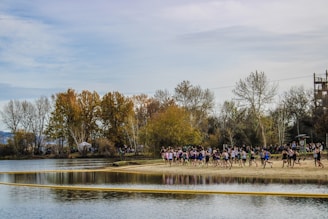 A group of people running along the sandy edge of a lake surrounded by autumn trees. The sky is overcast, and the water reflects the surrounding landscape.