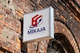 A sign with the text 'MIKAJA' and 'Agencia de envios a Cuba' attached to a brick wall. The sign features a stylized logo with a red and white box, a blue arrow, and a white star on a red background. The brick wall has several patches, some exposed wires, and a partially visible arched window.