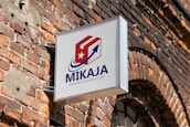 A sign with the text 'MIKAJA' and 'Agencia de envios a Cuba' attached to a brick wall. The sign features a stylized logo with a red and white box, a blue arrow, and a white star on a red background. The brick wall has several patches, some exposed wires, and a partially visible arched window.