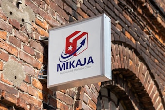 A sign with the text 'MIKAJA' and 'Agencia de envios a Cuba' attached to a brick wall. The sign features a stylized logo with a red and white box, a blue arrow, and a white star on a red background. The brick wall has several patches, some exposed wires, and a partially visible arched window.