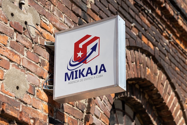 A sign with the text 'MIKAJA' and 'Agencia de envios a Cuba' attached to a brick wall. The sign features a stylized logo with a red and white box, a blue arrow, and a white star on a red background. The brick wall has several patches, some exposed wires, and a partially visible arched window.