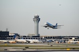 An aircraft is taking off from an airport runway, with another airplane stationary in the background. The airport control tower is visible behind them, along with terminal buildings and some ground vehicles. The sky is clear with a small plane flying high further in the background.