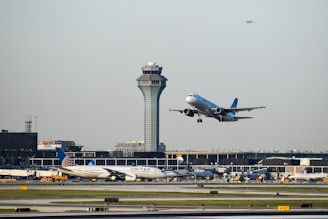 An aircraft is taking off from an airport runway, with another airplane stationary in the background. The airport control tower is visible behind them, along with terminal buildings and some ground vehicles. The sky is clear with a small plane flying high further in the background.