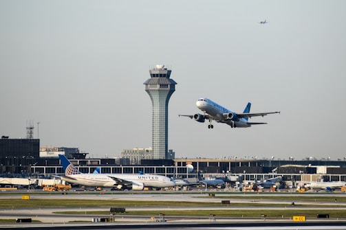 An aircraft is taking off from an airport runway, with another airplane stationary in the background. The airport control tower is visible behind them, along with terminal buildings and some ground vehicles. The sky is clear with a small plane flying high further in the background.