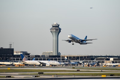 An aircraft is taking off from an airport runway, with another airplane stationary in the background. The airport control tower is visible behind them, along with terminal buildings and some ground vehicles. The sky is clear with a small plane flying high further in the background.