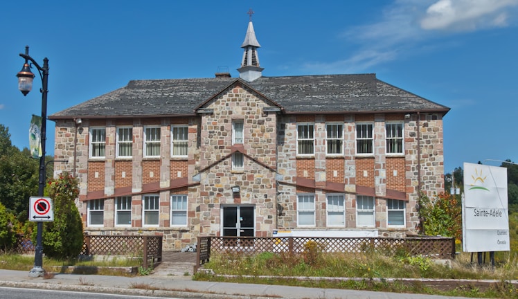 A two-story stone building with a steeply pitched roof and numerous windows. The structure has a sign in front indicating 'Sainte-Adele, Quebec, Canada.' A small steeple is present on the roof. A lamppost and a no-parking sign are situated along the sidewalk.