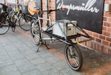 A cargo bicycle is parked on a paved sidewalk outside a building with brick walls. It has a large metal frame designed for carrying goods, with a gray storage box securely placed on its front. Other bicycles are parked nearby, and a sign with text is visible on the wall behind.