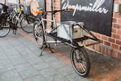A cargo bicycle is parked on a paved sidewalk outside a building with brick walls. It has a large metal frame designed for carrying goods, with a gray storage box securely placed on its front. Other bicycles are parked nearby, and a sign with text is visible on the wall behind.