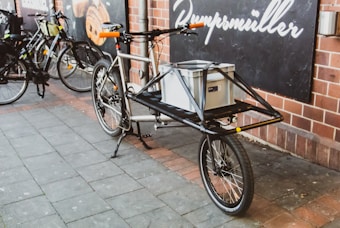 A cargo bicycle is parked on a paved sidewalk outside a building with brick walls. It has a large metal frame designed for carrying goods, with a gray storage box securely placed on its front. Other bicycles are parked nearby, and a sign with text is visible on the wall behind.