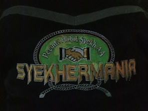 An emblem depicting two hands shaking in unity above the text 'Syehkermania'. The background includes a green circular shape bordered by a rope-like pattern, with the words 'Pecinta Habib Syekh AA' above the clasping hands.