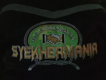 An emblem depicting two hands shaking in unity above the text 'Syehkermania'. The background includes a green circular shape bordered by a rope-like pattern, with the words 'Pecinta Habib Syekh AA' above the clasping hands.