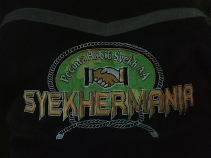 An emblem depicting two hands shaking in unity above the text 'Syehkermania'. The background includes a green circular shape bordered by a rope-like pattern, with the words 'Pecinta Habib Syekh AA' above the clasping hands.