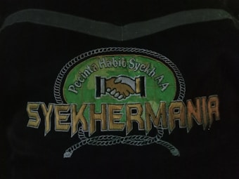 An emblem depicting two hands shaking in unity above the text 'Syehkermania'. The background includes a green circular shape bordered by a rope-like pattern, with the words 'Pecinta Habib Syekh AA' above the clasping hands.