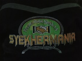 An emblem depicting two hands shaking in unity above the text 'Syehkermania'. The background includes a green circular shape bordered by a rope-like pattern, with the words 'Pecinta Habib Syekh AA' above the clasping hands.