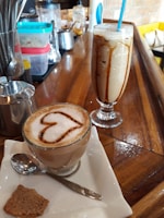 A glass mug of coffee with frothy milk is topped with a heart-shaped cinnamon or chocolate design, accompanied by a spoon and a piece of biscuit on a napkin. Beside it, there is a tall glass of iced coffee with a blue straw, drizzled with syrup. The wooden countertop displays various containers and utensils in the background.