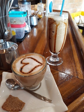 A glass mug of coffee with frothy milk is topped with a heart-shaped cinnamon or chocolate design, accompanied by a spoon and a piece of biscuit on a napkin. Beside it, there is a tall glass of iced coffee with a blue straw, drizzled with syrup. The wooden countertop displays various containers and utensils in the background.