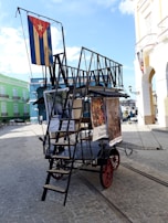 A wheeled cart with a metal frame and red rims is positioned on a cobblestone street. The cart is adorned with a large vertical ladder and a couple of posters or banners featuring artwork and text. A flag featuring a red star is prominently displayed on the cart, suggesting a cultural or artistic theme. The scene is set in a vibrant, colorful street with buildings painted in pastel shades of green, blue, and yellow. The sky is clear with a few scattered clouds.