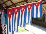 Multiple Cuban flags are hanging under a thatched roof structure. The flags are blue and white striped with red triangles featuring a white star. A collection of colorful shirts, including one with a notable face design, is visible to the left. A wooden hut and a fire extinguisher can be seen in the background.
