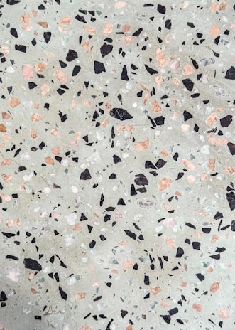 A terrazzo surface features an uneven distribution of multicolored stone chips embedded in a cementitious base. The chips are predominantly black, pink, and white, contrasting against a light grayish-green background.