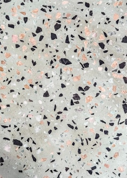 A terrazzo surface features an uneven distribution of multicolored stone chips embedded in a cementitious base. The chips are predominantly black, pink, and white, contrasting against a light grayish-green background.