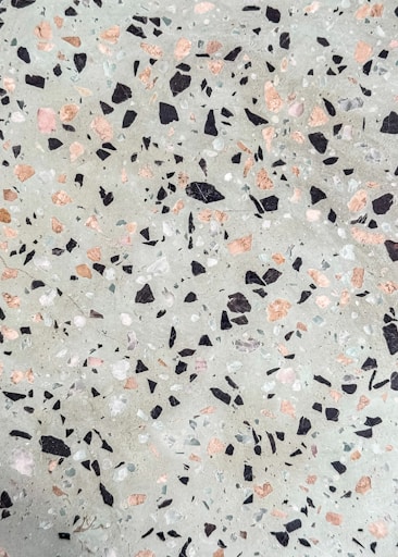 A terrazzo surface features an uneven distribution of multicolored stone chips embedded in a cementitious base. The chips are predominantly black, pink, and white, contrasting against a light grayish-green background.